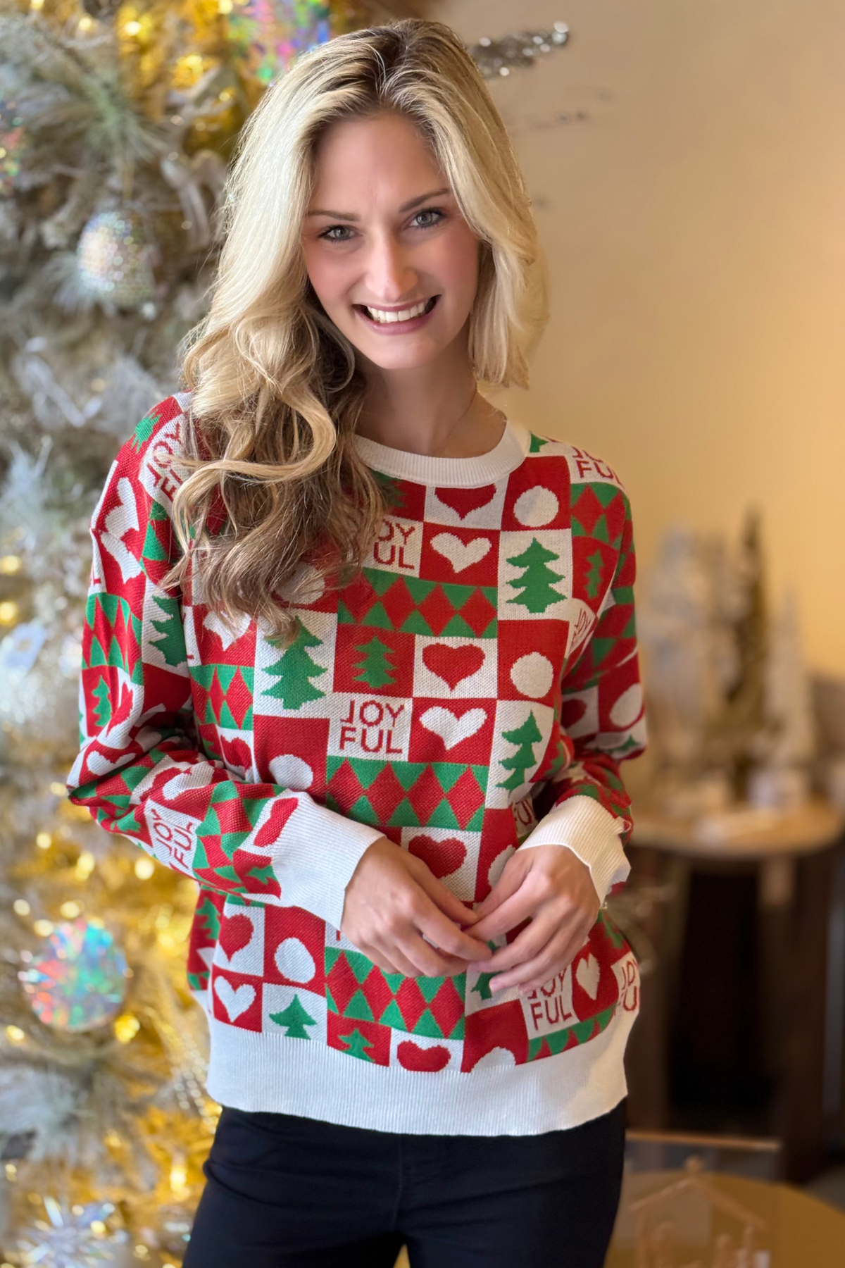 Festive holiday sweater with Christmas tree and heart print modeled indoors by a Christmas tree.