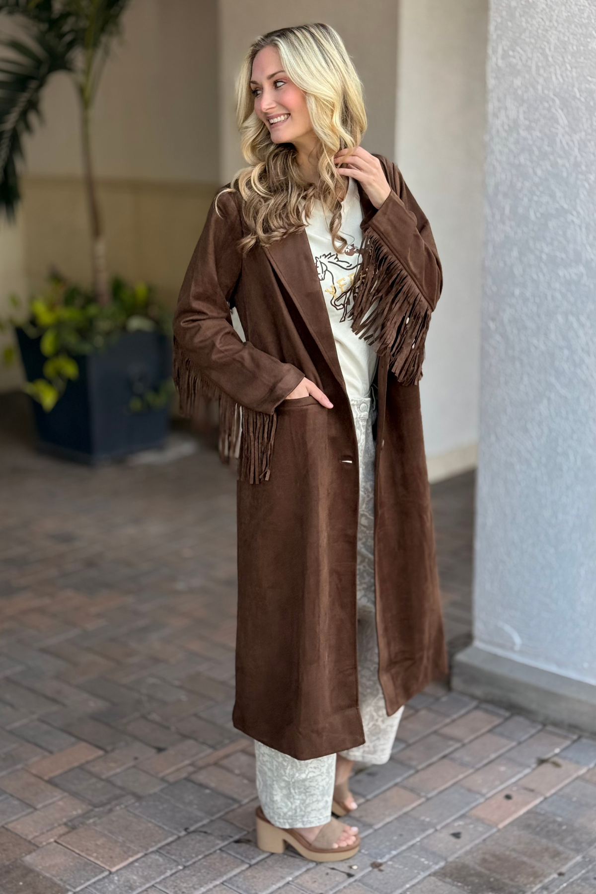 Woman wearing a brown fringed coat standing on a paved walkway.