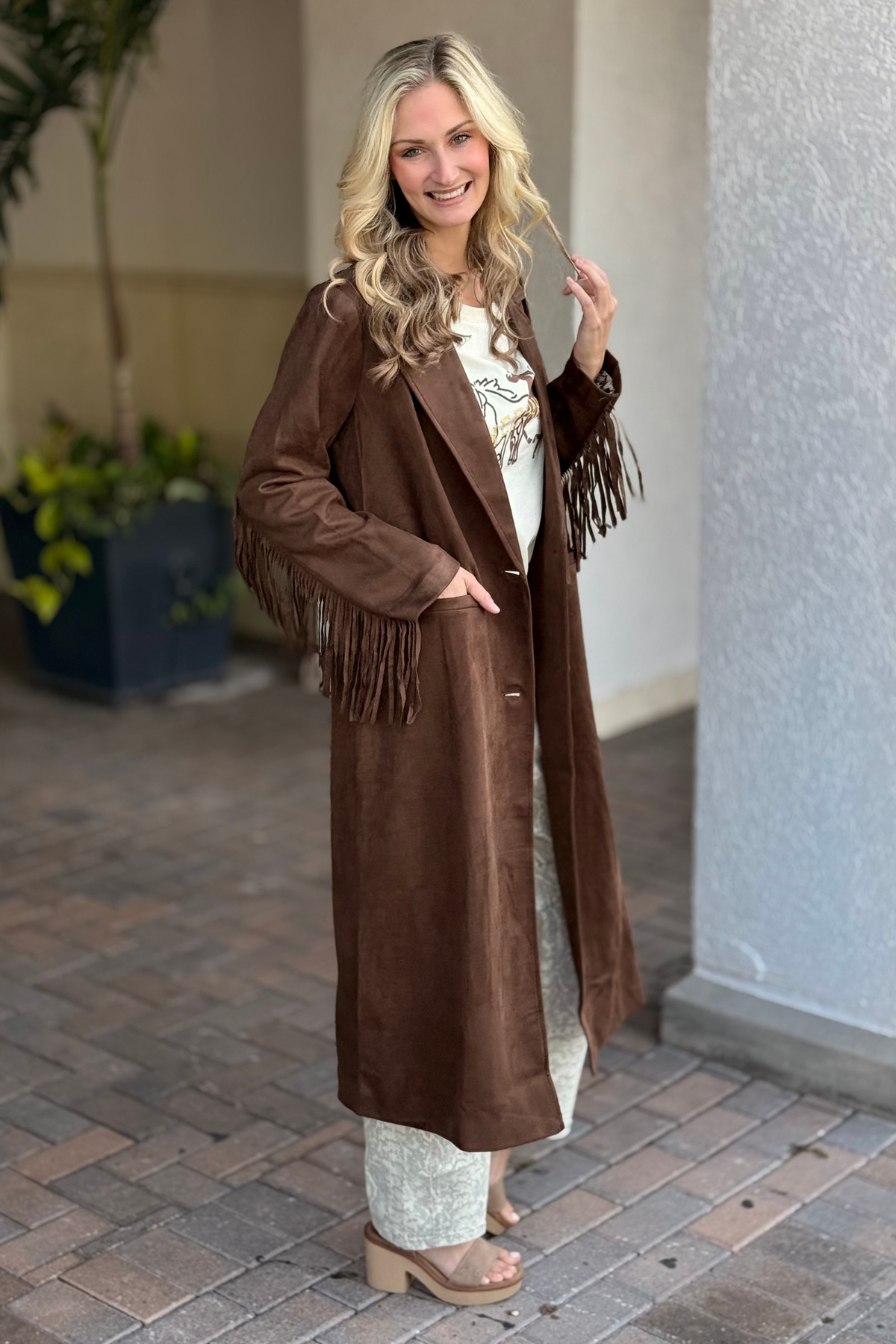 Woman wearing a long brown coat with fringe standing on a paved walkway.