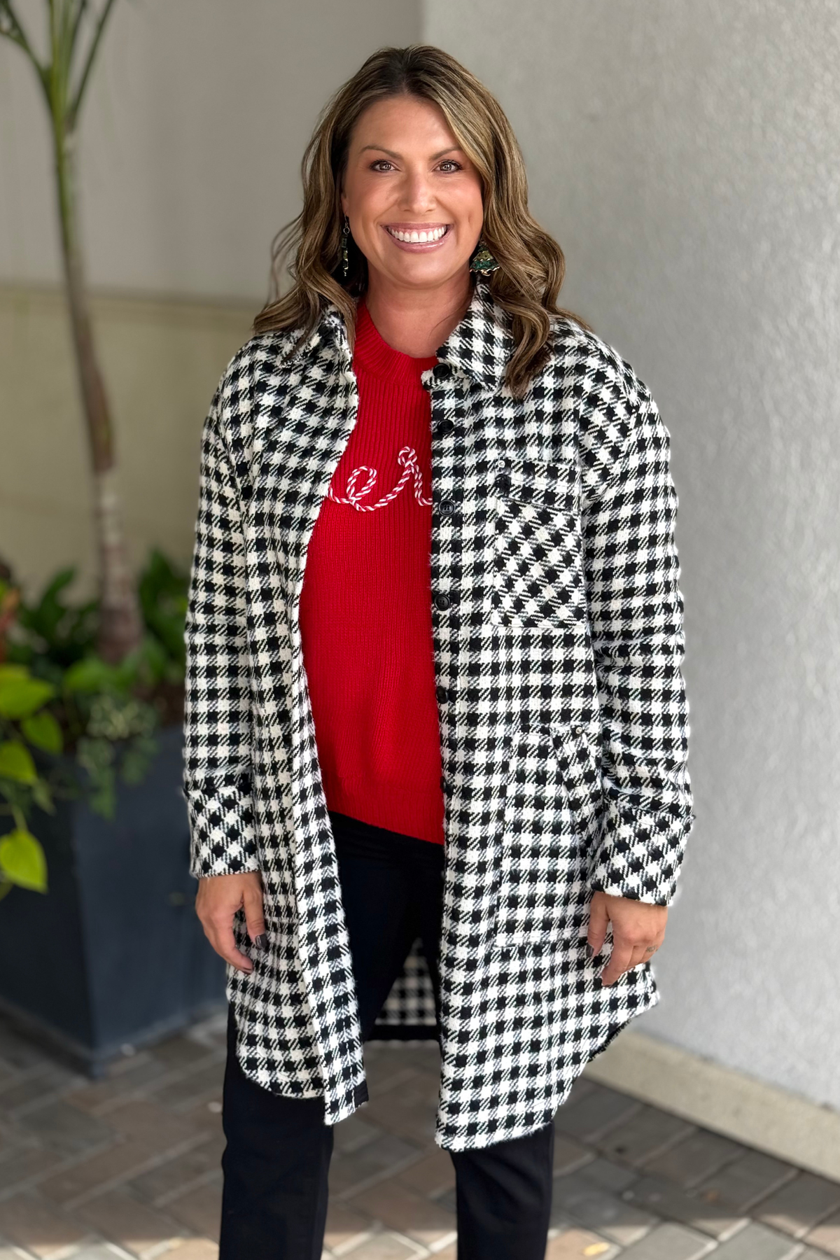 Woman wearing a black and white houndstooth longline shacket over a red “Merry” sweater, standing outdoors.