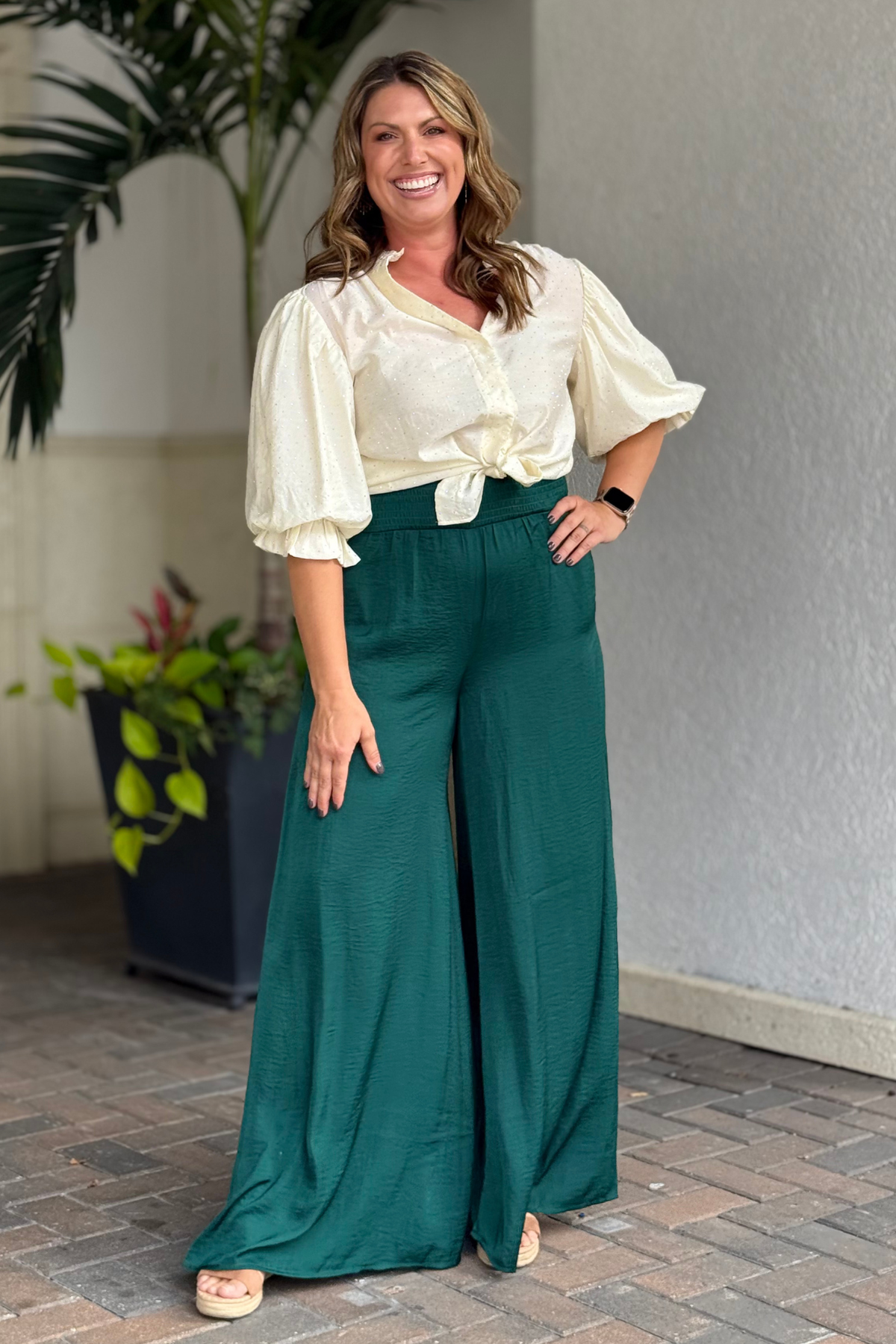 Woman wearing emerald green wide-leg satin pants with smocked waistband, styled with cream blouse