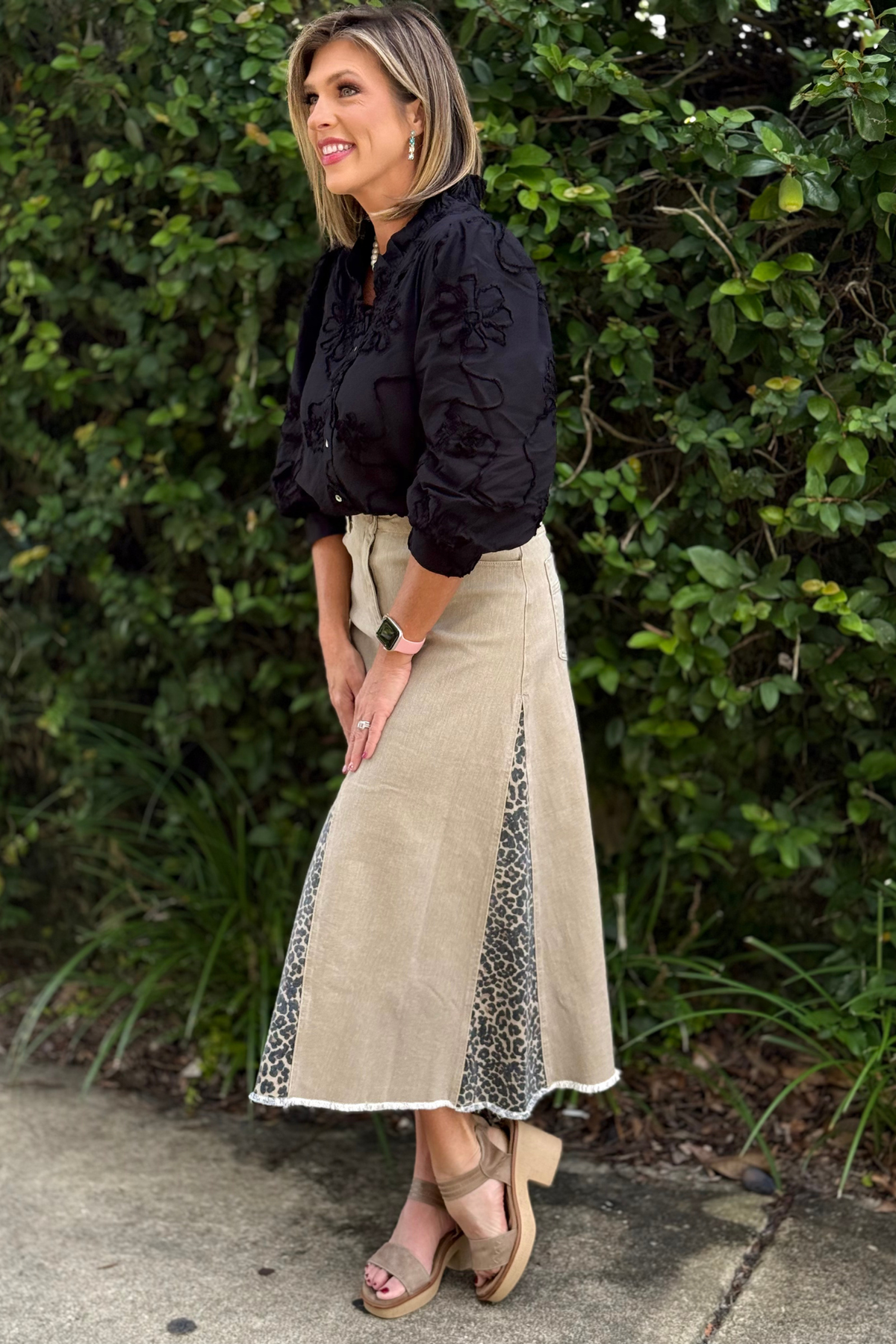 Woman wearing The Harper Panel Maxi Skirt — khaki maxi skirt with leopard print panels and frayed hem, styled with a black blouse and platform sandals.
