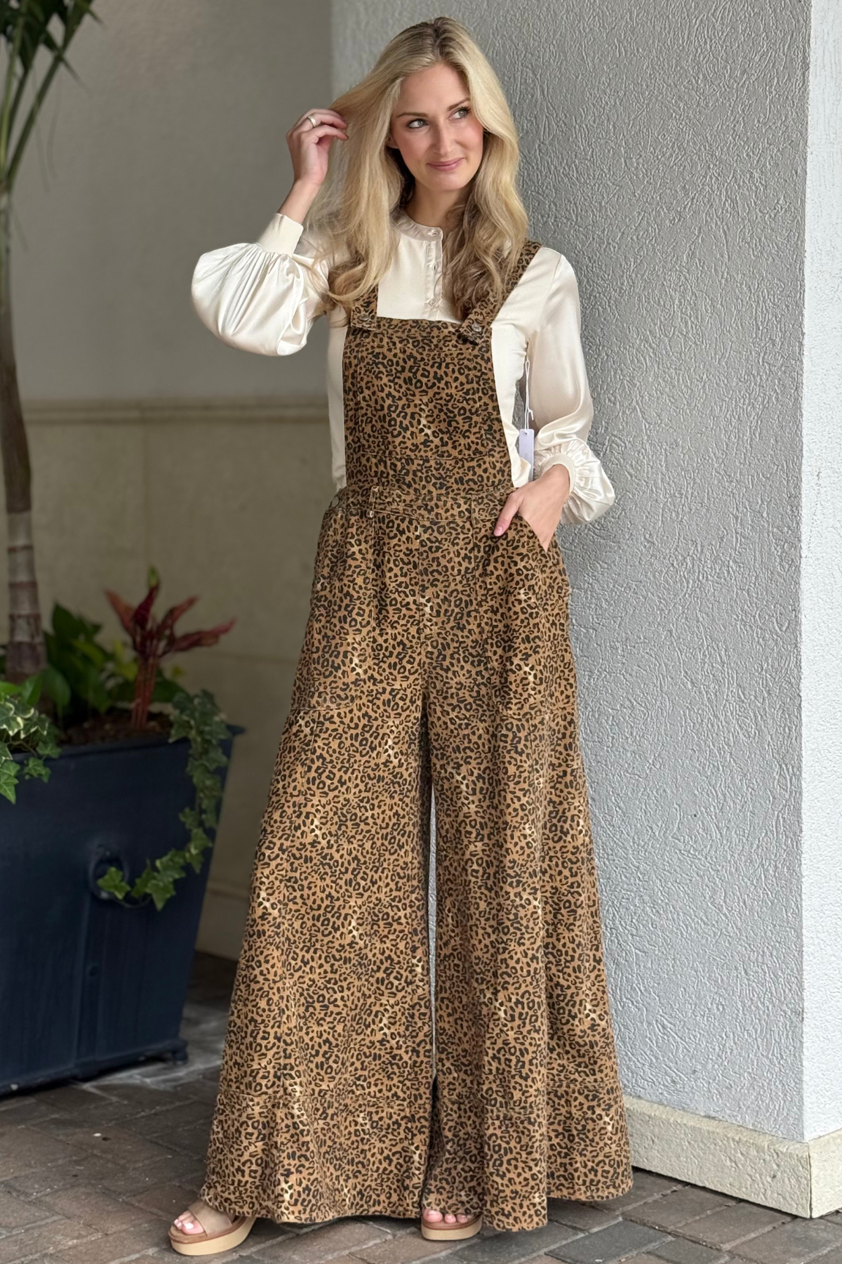 Woman wearing The Dakota Leopard Wide Leg Overalls — brown animal print overalls with wide legs and adjustable straps, styled with a satin blouse and platform sandals.