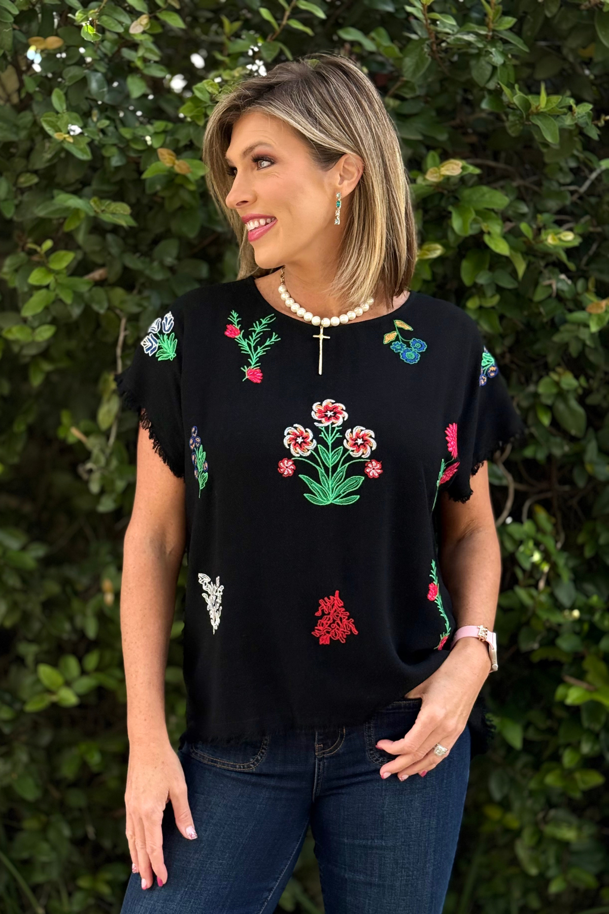 Woman wearing a black linen-blend embroidered top with colorful floral detailing, raw-edge sleeves, and frayed hemline, styled with denim jeans.