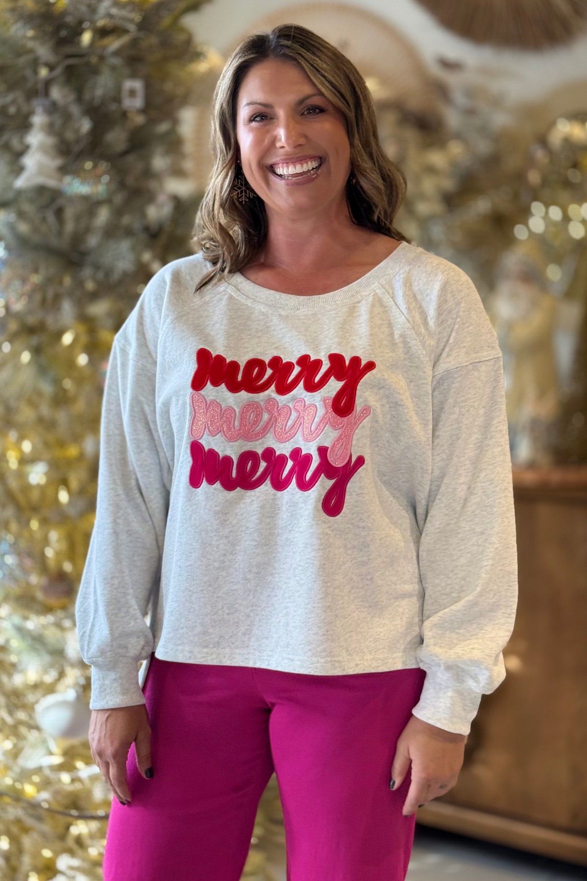 Woman wearing light heather gray sweatshirt with red and pink embroidered “merry” lettering and high-low hem, holiday outfit