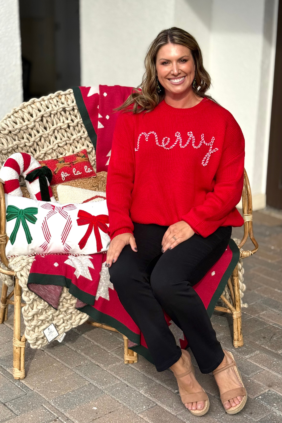 Woman wearing a red knit sweater with “Merry” embroidered in candy-cane design, standing by a decorated Christmas tree.