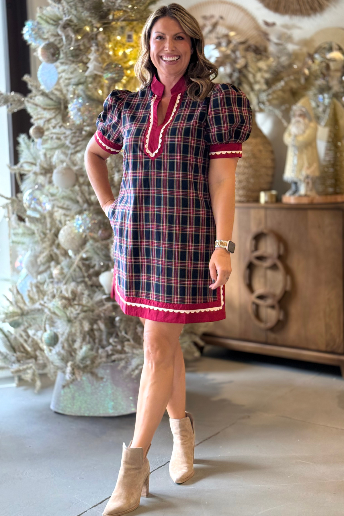 Woman wearing red and green plaid mini dress with scalloped trim and puff sleeves, styled with tan ankle boots.