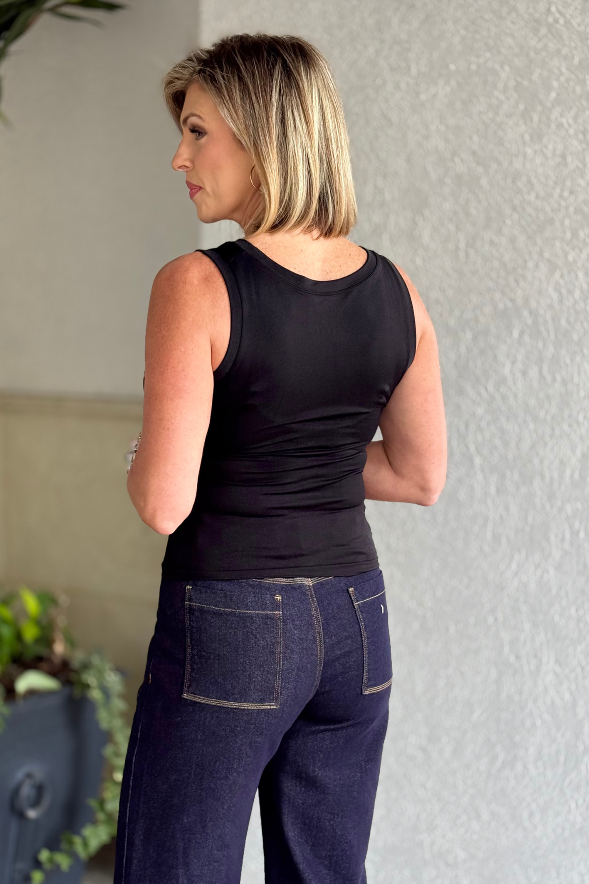 Woman wearing a black tank top and dark jeans standing against a light-colored wall.