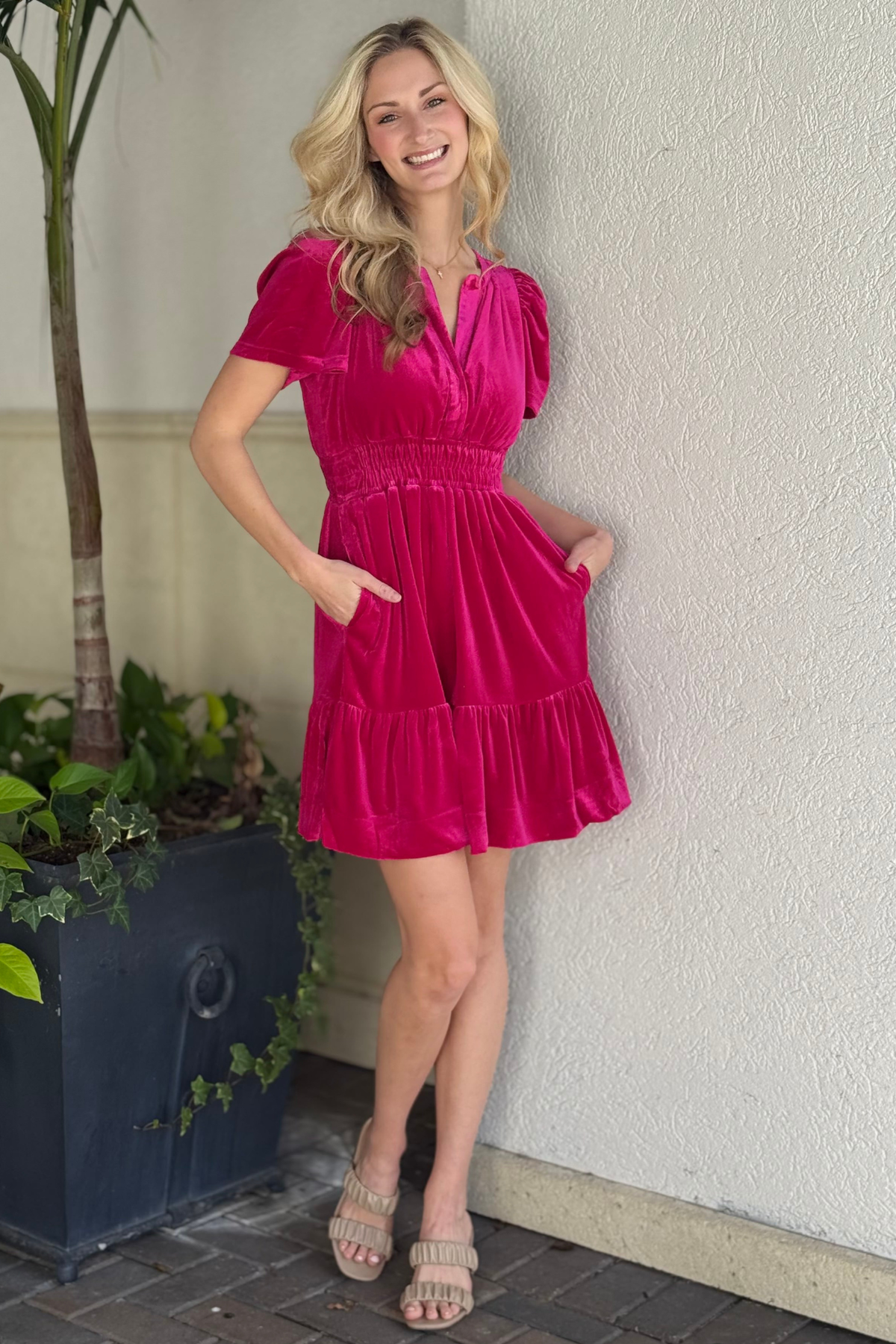 THML short sleeve velvet dress with pockets. Woman in a fuchsia dress standing against a light-colored wall with plants in the background.