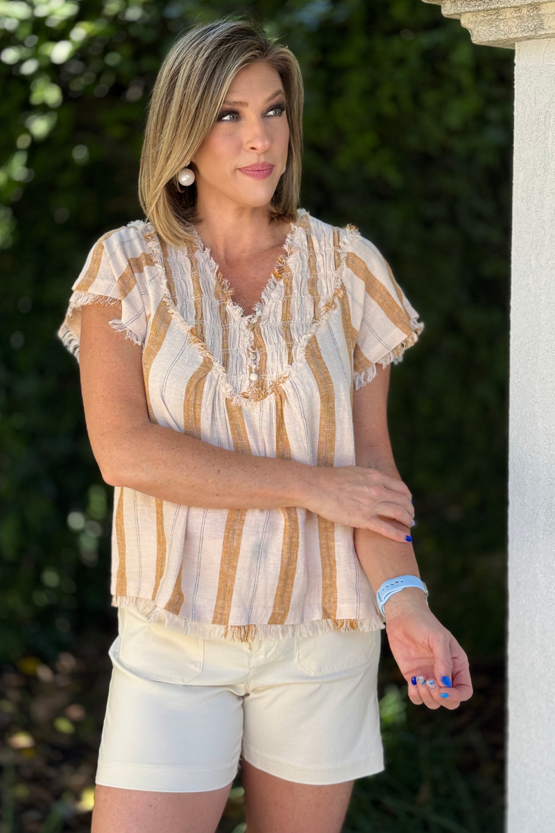 Woman wearing neutral striped linen blouse with smocked V neckline and cream shorts