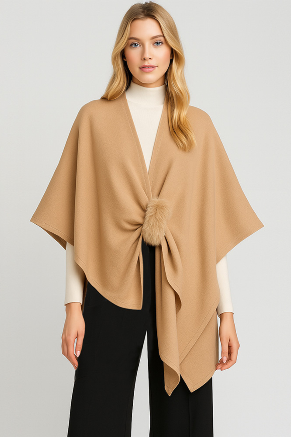 Woman wearing a camel knit ruana with faux fur pom closure, soft draped open-front poncho.