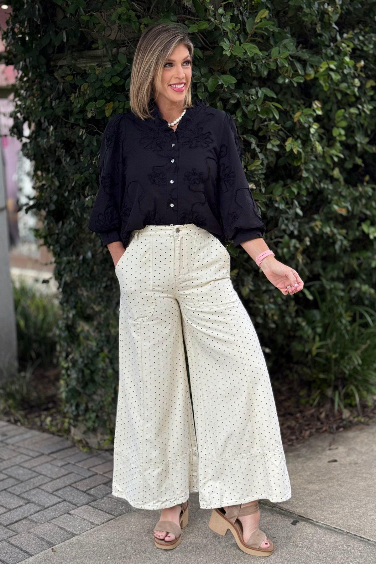 Woman wearing ivory wide-leg pants with black polka dots, high-rise waist, and front pockets, styled with a black textured blouse and wedge sandals outdoors.