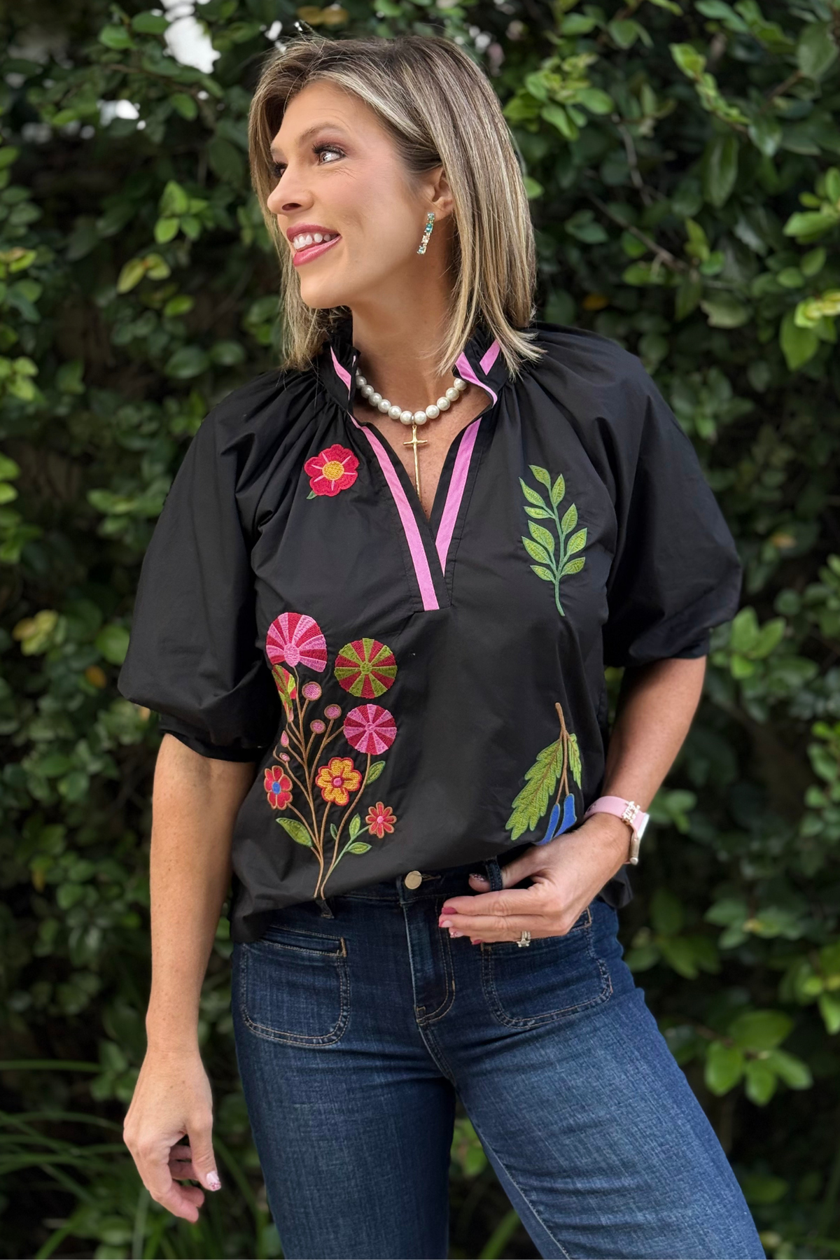 Woman wearing The Valencia Embroidered Blouse — black V-neck top with pink trim, colorful floral embroidery, and puff sleeves.