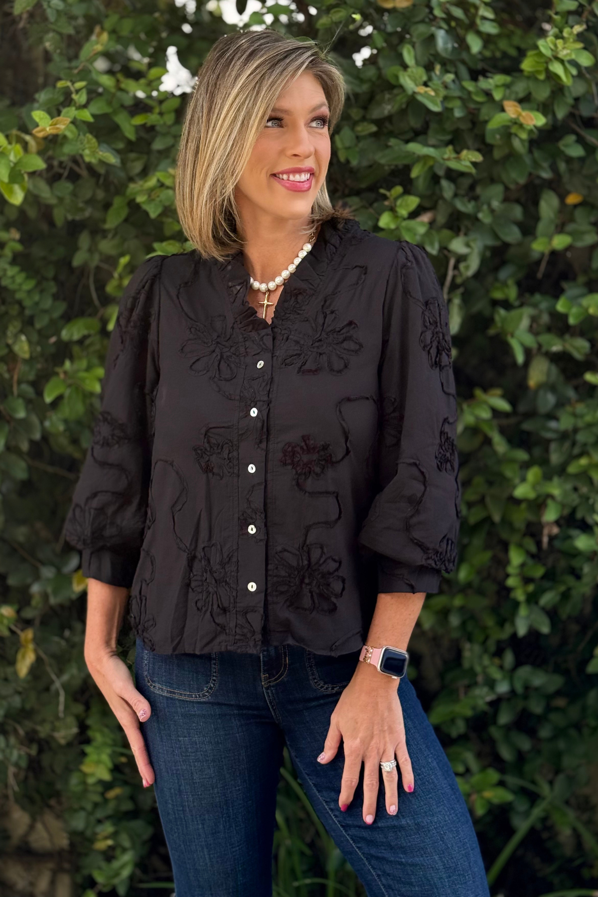 Woman wearing a black 3D floral textured blouse with 3/4 sleeves, ruffled neckline, and button-down front, styled with dark denim jeans, standing in front of greenery.