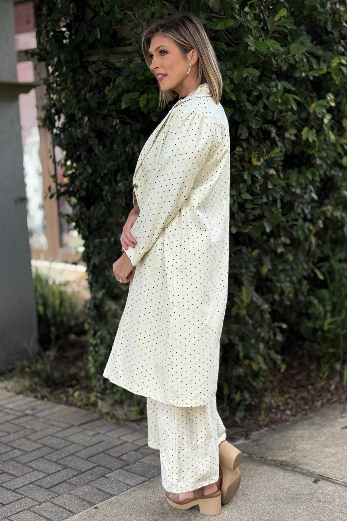 Woman wearing a cream-colored polka dot longline duster jacket with a tailored collar, button front, and side pockets, styled over matching wide-leg pants and a black blouse.