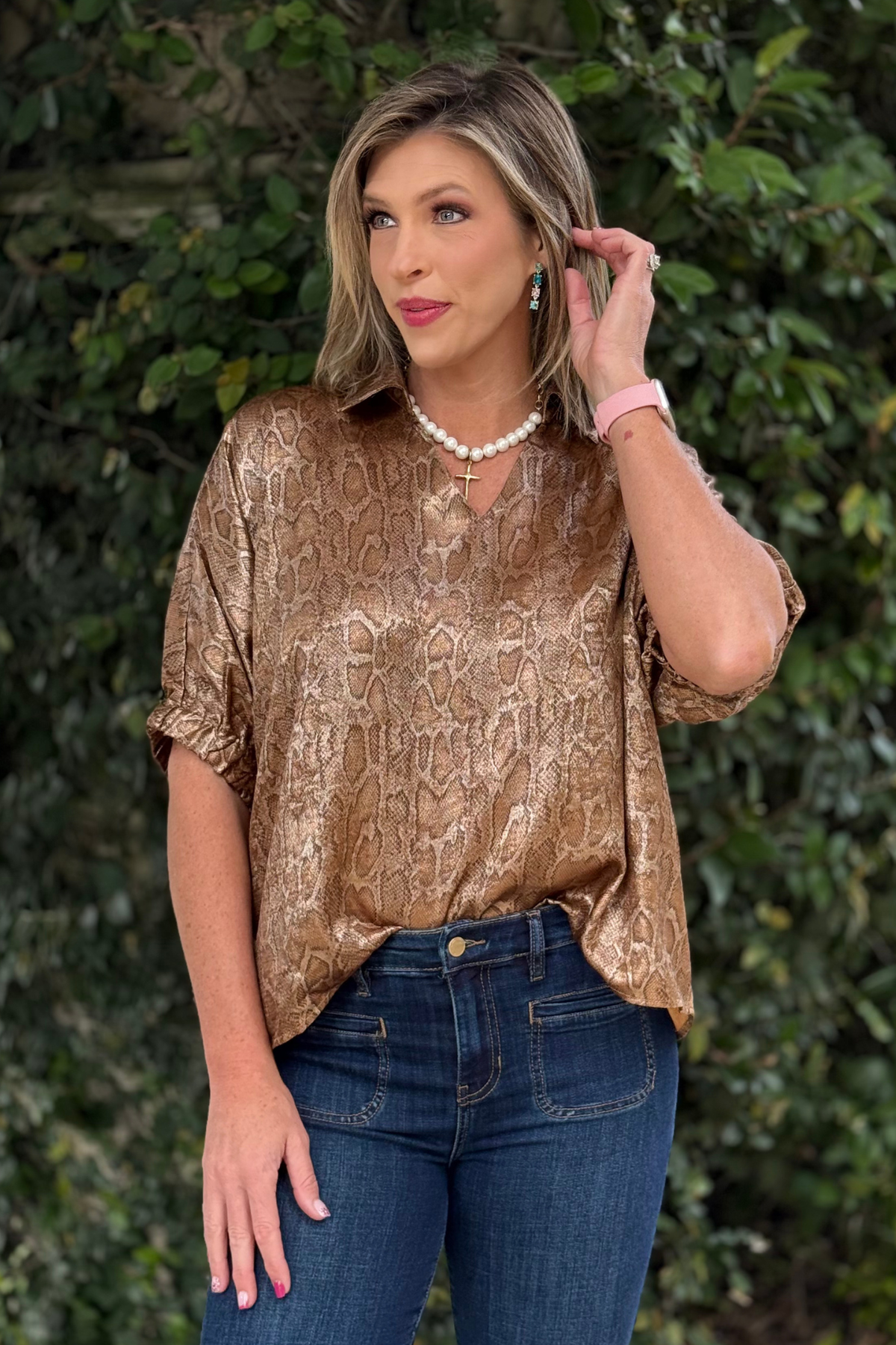 Woman wearing a gold snakeskin shimmer top with collared v-neckline, elastic short sleeves, and a subtle sheen, styled with dark denim jeans.